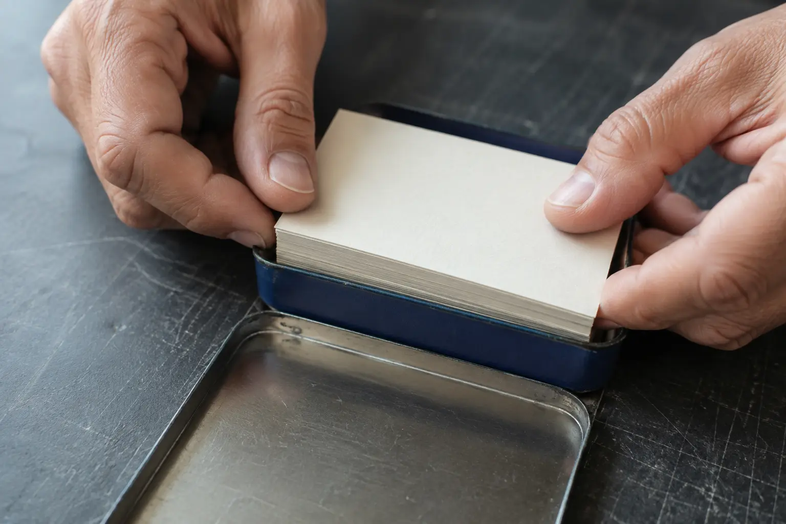 Adult hands placing a compact blank card stack into a plain blue metal pocket tin